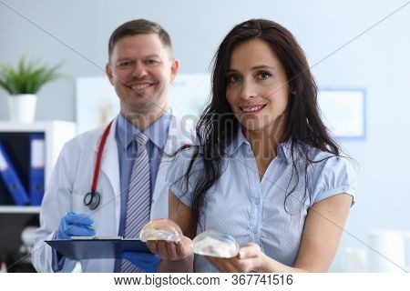 Girl In Clinic Holds Breast Implants Near Doctor. Comfortable Atmosphere. Work Plastic Surgeon. Sele