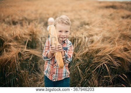 A Blond Boy In An Orange Plaid Shirt And Green Trousers Stands In A Rye Field, Smiles And Holds A Wh