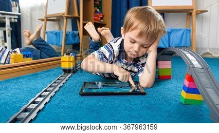 Cute Little Boy Lying On Floor And Playing Educational Games On Tablet Computer