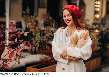 Portrait Of A Pretty Woman In A Red Beret And A White Cardigan With Baguettes In Her Hands Strolling