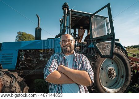 Portrait Of Satisfied Tractor Driver After Work On Agricultural Field Stands Next To Tractor. Farmin
