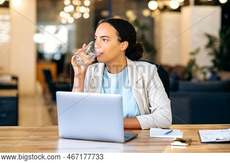 Healthy Brazilian Or Hispanic Business Lady, Sits At A Workplace In The Office, Drinking A Glass Of 