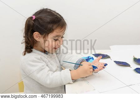 Little Girl Painting Leaves Blue Color, Crafts And Art Therapy. Child Paints Her Own Hand With Blue 