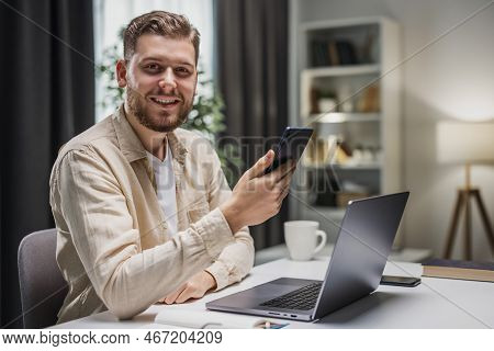 Young Handsome Man Sitting At Desk With Aptop, Holding Smartphone And Smiling To Camera.