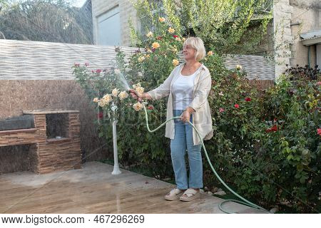 Portrait Of Smiling Senior Woman Holding Water Hose Watering Flowers At Backyard Garden. Growing And