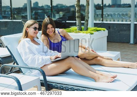 Two Beautiful Young Female Friends In Swimsuits Are Lying On Deckchairs By The Pool And Looking At I