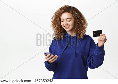 Portrait Of Curly Girl Smiles, Makes Order, Shops Online In App, Shows Credit Card And Holds Mobile 