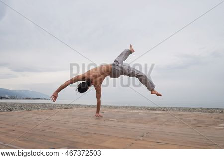 Shirtless Caucasian Man Doing Backflip On Pebble Beach.
