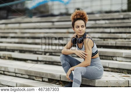 A Serious Runner In Sportswear, With Headphones Around Her Neck, Sits On The Stairs Outdoors And Tak