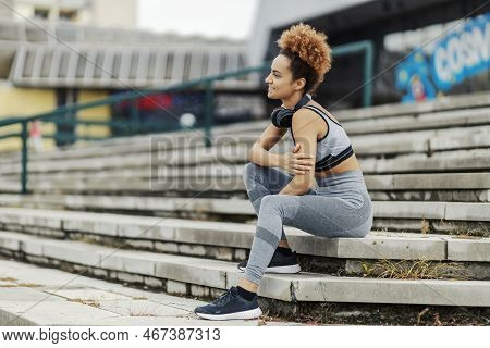 A Happy Sportswoman In Sportswear, With Headphones Around Her Neck, Sits On The Stairs Outdoors In T