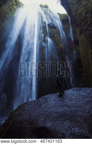 Man Near High Waterfall Landscape Photo. Beautiful Nature Scenery Photography With Cliffs On Backgro