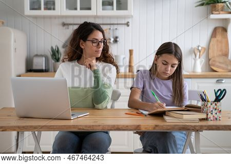 Work-at-home Parent Mother Looking At Daughter Doing Homework, Sit Together At Table In Kitchen. Sch