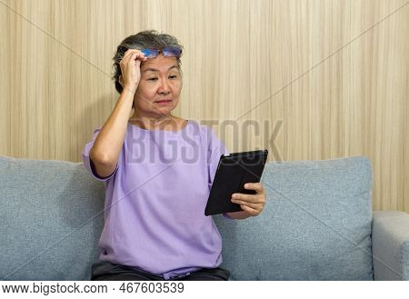 Asian Retired Elderly With Eyeglasses, Sitting On The Sofa Watching At Tablet Computer. With Presbyo