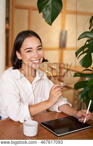 Beautiful Asian Girl Sitting In Cafe With Cup Of Coffee, Drawing On Digital Tablet With Graphic Pen,