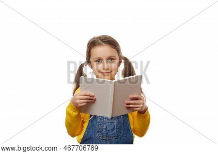 Smart First Grade Schoolgirl, With Two Ponytails, Wearing A Yellow Pull And Denim Overalls, Reading 