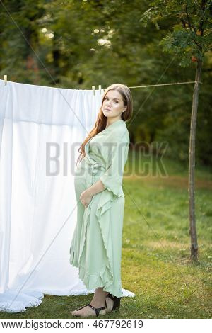 Beautiful Pregnant Woman With Big Belly Stands Near Clothesline With Bed Sheet, Young Female Takes C