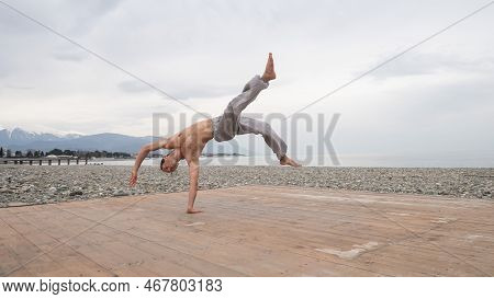 Shirtless Caucasian Man Doing Backflip On Pebble Beach.