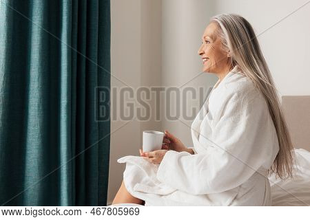 Senior Woman Sitting On Bed In Bedroom And Looking Away. Side View Of Pensive Mature Female.