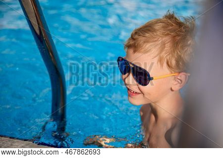 Smiling Boy In Sunglasses Swam Up To The Steps In The Pool To Climb. Happy Child Swims In The Warm C