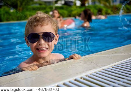 Cheerful Child In Sunglasses Smiles, Swims In The Warm Clear Water In The Pool. Fashionable Child In
