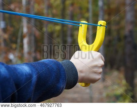 Slingshot With A Stretched Bowstring In A Female Hand Against The Backdrop Of A Forest