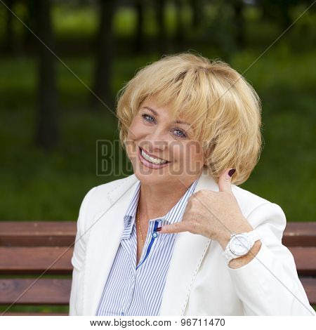 Happy smiling old blonde woman with call me gesture, against green sunner park