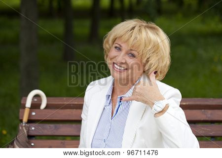 Happy smiling old blonde woman with call me gesture, against green sunner park
