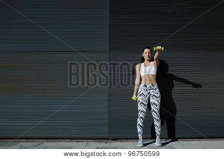 Athletic young woman lifting weights standing with arms tense against black wall outdoors