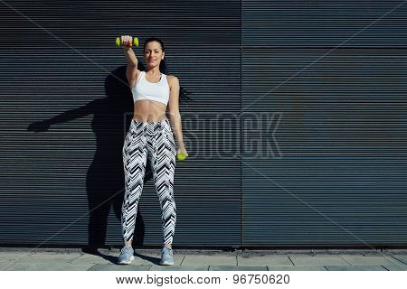 Sporty young woman lifting weights standing with arms tense against black wall outdoors
