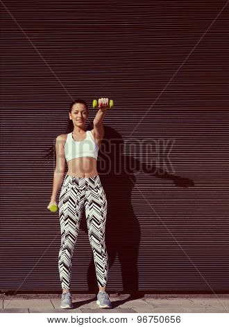 Smiling young woman lifting weights standing with arms tense against black wall background at sunset