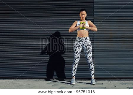 Confident sporty woman using dumbbells to work out her arms while training outdoors