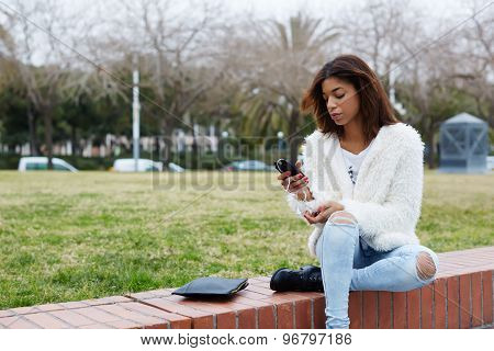 Portrait of young stylish hipster girl holding smart phone while sitting in the park outdoors