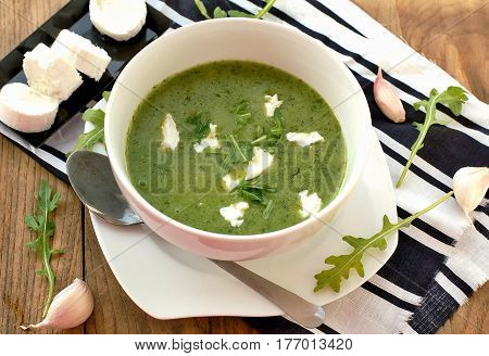Green Soup From Arugula And Garlic With Goat Cheese In White Bowl With Spoon On Wooden Background