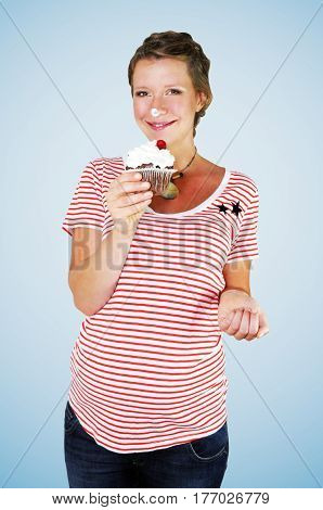 Smiling pregnant young woman wearing and red and white striped oversized T-shirt and maternity jeans is snacking on a large chocolate muffin with extra whipped cream and a cherry on top on blue background.
