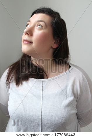 Portrait of pale brunette, green eyed Caucasian woman in her twenties looking up at light coming in through a high window. Wearing a white top, lipstick and blusher and against a white wall