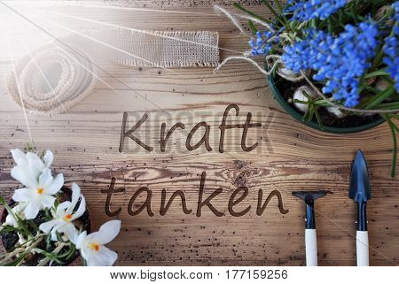 German Text Kraft Tanken Means Relax. Sunny Spring Flowers Like Grape Hyacinth And Crocus. Gardening Tools Like Rake And Shovel. Hemp Fabric Ribbon. Aged Wooden Background