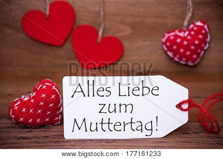 Label With German Text Alles Liebe Zum Muttertag Means Happy Mothers Day. White Label With Red Textile Hearts. Retro Brown Wooden Background.