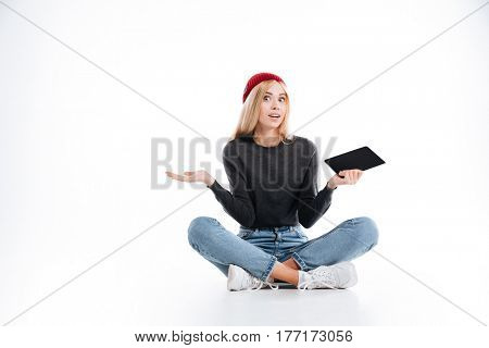 Uncomprehending female hipster which sitting on the floor in studio with tablet computer and looking at camera over white background