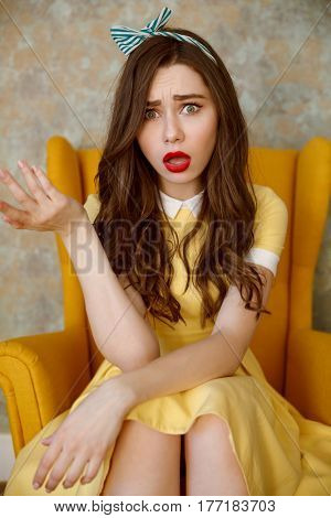 Vertical image of Uncomprehending woman in yellow dress which sitting on armchair and looking at camera