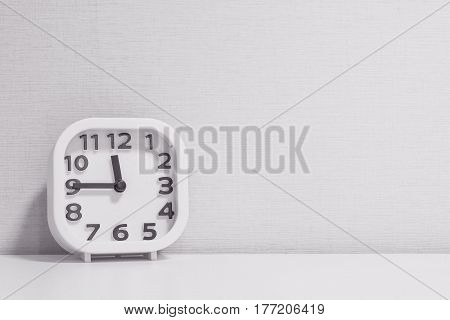 Closeup white clock for decorate show a quarter to twelve o'clock or 11:45 a.m. on white wood desk and cream wallpaper textured background in black and white tone with copy space