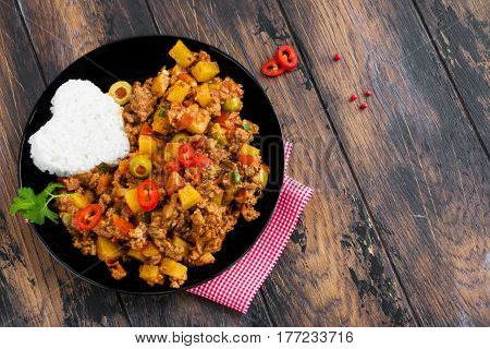 Picadillo a stew of ground beef and tomatoes with raisins and olives with boiled rice on a black plate. Wooden table top view.