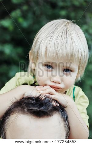 Tired cute baby boy son little child with blond hair sitting on father neck or shoulders on sunny summer day outdoors on natural background