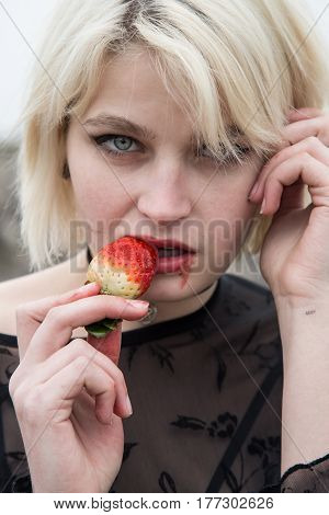 Beautiful young Russian girl with blond hair and blue eyes eating a strawberry in the field. She is dressed in a transparent black dress.