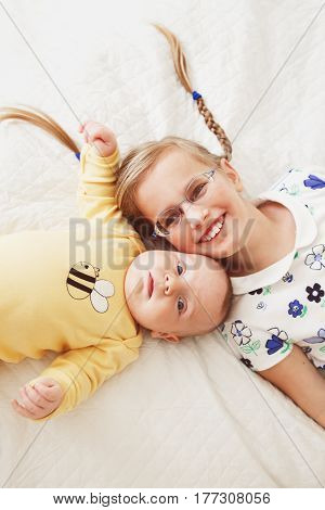Portrait of cute adorable little white Caucasian girl sister with braids and newborn baby sibling brother lying on bed in bedroom lifestyle real true emotions view from top above