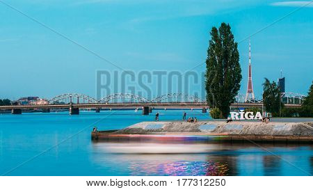 Riga, Latvia. The Concrete Embankment At Daugava River With City Name Sign And Resting People Around. Radio TV Tower, Stone And Railway Bridges Background In Summer Evening Dusk.