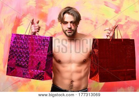 Bearded man short beard. Caucasian sexy macho man with moustache shows six packs and abs on muscle torso holding red packages or bags on colorful studio background