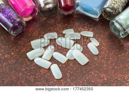 Artificial plastic nail near bottles of nail polish varnish lacquer, pieces of fake Acrylic nails collection on dark brown background 