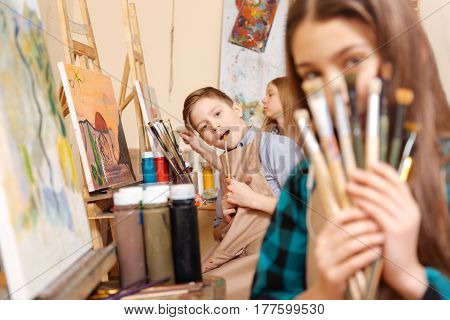 Having fun together. Playful joyful positive kids sitting in the art studio and having painting class while expressing joy and holding paintbrushes