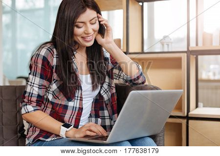 Age of technology. Delighted happy positive woman holding a laptop and looking at its screen while speaking with somebody on the phone