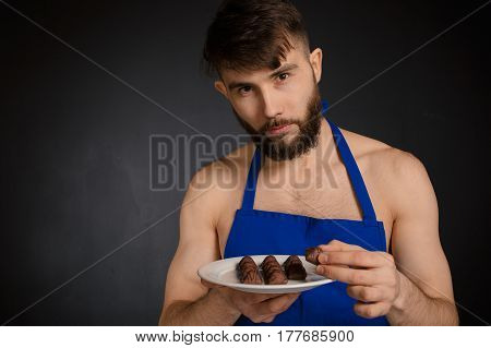 Hot sexy naked handsome man with chocolates chocolate candies. Smiling naked handsome man wearing dark blue apron holding a white plate with many sweets chocolate candies. Handsome confectioner.
